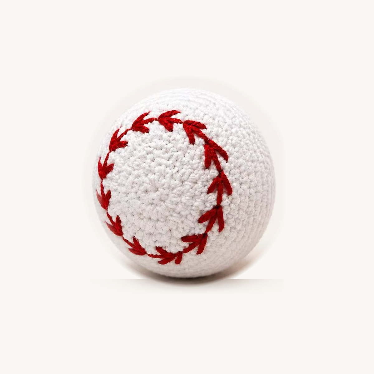 White crochet-style baseball dog toy with red stitching, on an ivory background; approximately 8 cm.
